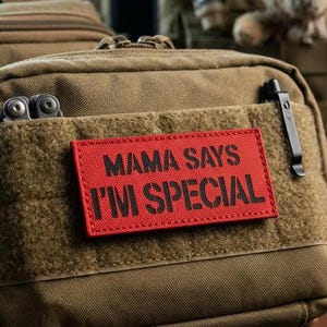 Pode incluir: Uma bolsa tática bege com um emblema retangular vermelho com o texto preto MAMA SAYS I'M SPECIAL. A bolsa possui um fecho de correr, um painel de velcro frontal e um clipe de metal preto preso na borda lateral do bolso frontal.