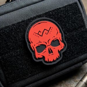 May include: A rectangular black tactical pouch featuring a red skull patch with black detailing on the front. The pouch has a zippered closure and a hook-and-loop panel. Beside the pouch lies a metallic pen and a black folding pocket knife.