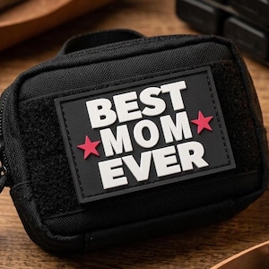 Peut inclure: Une pochette tactique rectangulaire noire dotée d'un écusson amovible avec le texte blanc "BEST MOM EVER" et deux petites étoiles rouges sur fond noir. La pochette possède une fermeture éclair et une structure en tissu robuste avec un panneau velcro.