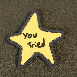 Puede incluir: Bolsa verde oliva con cremallera y parche en forma de estrella amarilla. El parche tiene las palabras "You Tried" en negro. La bolsa tiene una superficie texturizada.