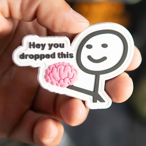 Puede incluir: Un parche de goma blanco con un monigote negro que sostiene un cerebro rosa. Un bocadillo junto a la figura contiene el texto negro "Hey you dropped this". El parche tiene un diseño moldeado en relieve con un borde blanco limpio.