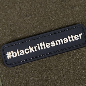 May include: Olive green zippered pouch with a textured surface. A black rectangular patch with the white text "#blackriflesmatter" is affixed to the front. The pouch has a loop and cord attached.