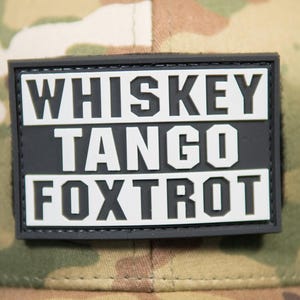 Może przedstawiać: Czapka bejsbolowa w kamuflażu z czarną siatką z tyłu. Przód czapki zdobi prostokątna naszywka z białym napisem "WHISKEY TANGO FOXTROT". Czapka posiada również naklejkę "MULTICAM" i metkę "SNAPBACK".