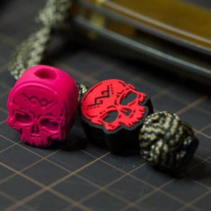 May include: Two skull-shaped beads, one red and one black, with a white skull design. The beads are strung on a cord with a black and gold braided cord.