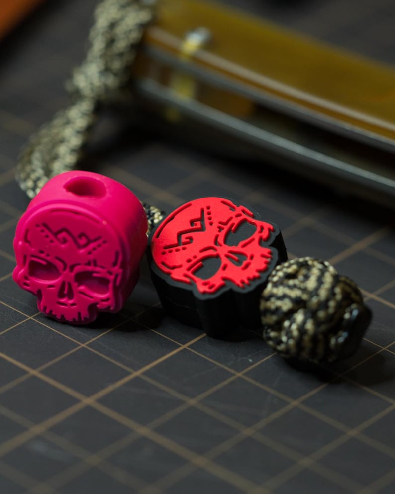 Puede incluir: Dos cuentas con forma de calavera, una roja y otra negra, con un dise&ntilde;o de calavera blanca. Las cuentas est&aacute;n enhebradas en un cord&oacute;n con un cord&oacute;n trenzado negro y dorado.