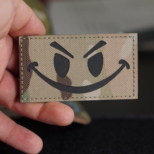 Op de afbeelding: Een bruine en tan camouflage stoffen patch met een zwart lachend gezicht met opgeheven wenkbrauwen. De patch is gestikt rond de randen.