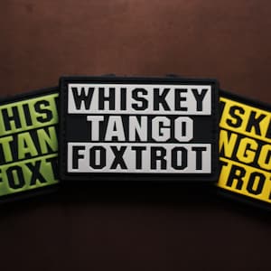 May include: Three rectangular patches with the text "WHISKEY TANGO FOXTROT" in bold, capital letters. The patches are black with white lettering, green with black lettering, and yellow with black lettering. The background is a dark brown.