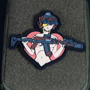 Pode incluir: Um emblema moral de PVC com uma ilustração estilizada de uma figura usando um capacete tático com óculos de visão noturna, segurando um rifle sobre o peito. O design é definido contra um fundo de coração rosa com borda preta.