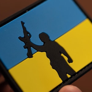 May include: A black patch featuring a silhouette of a person holding a rifle in front of the Ukrainian flag. The flag is blue at the top and yellow at the bottom.