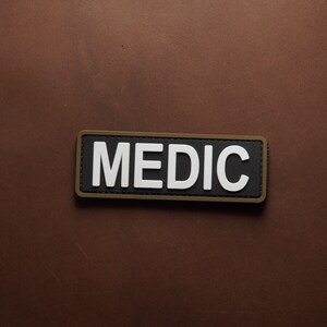 Pu&ograve; includere: Una toppa nera e bianca con la parola "MEDIC" in lettere bianche su sfondo nero. La toppa ha un bordo marrone.