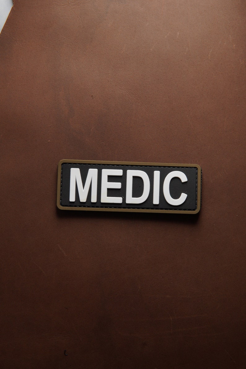 Pu&ograve; includere: Una toppa nera e bianca con la parola "MEDIC" in lettere bianche su sfondo nero. La toppa ha un bordo marrone.