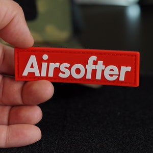 May include: A red rectangular patch with white stitching around the edges. The patch has the word "Airsoffter" printed in white letters.