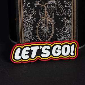 Könnte beinhalten: Ein rechteckiger Aufnäher mit schwarzem Hintergrund und einer weißen Strichzeichnung einer Person auf einem Fahrrad. Unter der Illustration befindet sich ein mehrfarbiges Schild mit dem weißen Schriftzug LET'S GO! und schwarzen, gelben und roten Rändern.