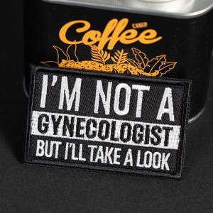 May include: A rectangular black embroidered patch featuring white stitched text that reads I'M NOT A GYNECOLOGIST BUT I'LL TAKE A LOOK. The patch has a thick black border and is positioned in front of a black container with yellow graphic details.