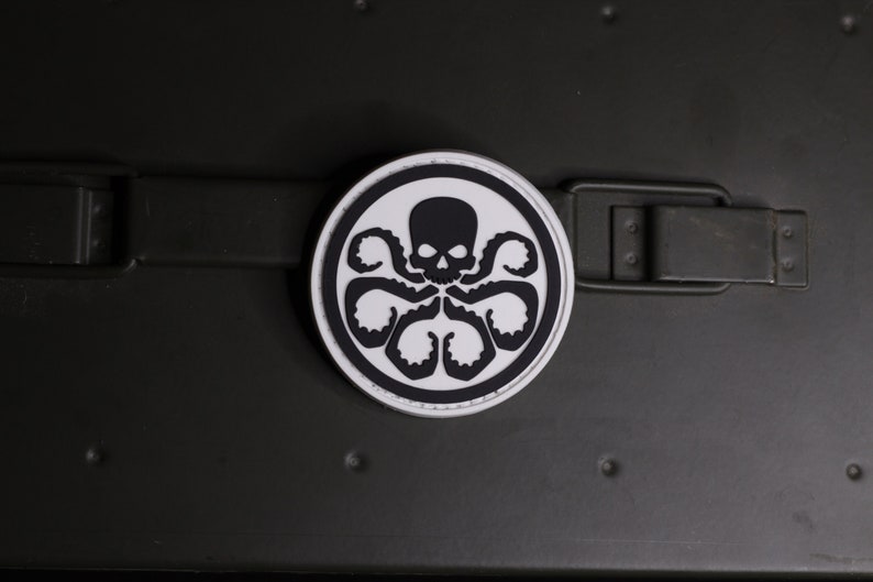 May include: A circular patch featuring a black graphic of a skull with six tentacles extending downwards, set against a white background with a thick black border. The design is centered on a dark olive green strap with a rectangular metal buckle.