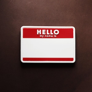 Könnte beinhalten: Ein weißer Namensschild mit rotem oberen Rand. Der Text "HELLO my name is" ist auf dem oberen Rand gedruckt.
