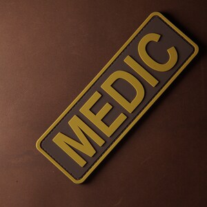 Pu&ograve; includere: Un cerotto rettangolare marrone con un bordo giallo e la parola "MEDIC" scritta in lettere gialle.