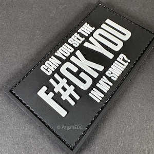 May include: Black rectangular patch with white text that reads "CAN YOU SEE THE F#CK YOU IN MY SMILE?". The patch has a stitched border and the text is in a bold, sans-serif font. The bottom left corner has the text "PaganEDC".