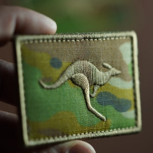 Può includere: Un cerotto in tessuto mimetico verde, marrone e nero con una silhouette di canguro ricamata in oro.