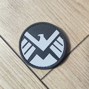 Puede incluir: Un parche circular con un fondo gris oscuro y un emblema de &aacute;guila gris claro centrado en la superficie. El dise&ntilde;o del &aacute;guila presenta alas estilizadas y un pico afilado, creando un contraste gr&aacute;fico audaz dentro del borde redondo del parche.