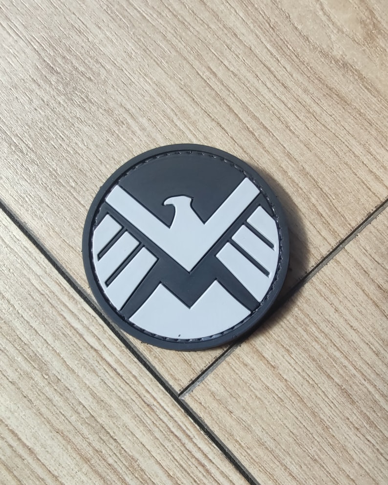 Puede incluir: Un parche circular con un fondo gris oscuro y un emblema de &aacute;guila gris claro centrado en la superficie. El dise&ntilde;o del &aacute;guila presenta alas estilizadas y un pico afilado, creando un contraste gr&aacute;fico audaz dentro del borde redondo del parche.