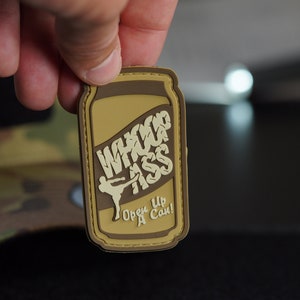 May include: A brown and tan embroidered patch shaped like a can of soda. The can has the words "Whup Ass" written on it in a stylized font. The can also has the words "Open Up A Can!" written on it.