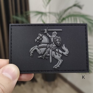 Puede incluir: Un parche rectangular negro con una imagen en relieve gris de un caballero a caballo con una espada y un escudo. El parche tiene una superficie texturizada y un borde. La letra "K" es visible en la esquina inferior derecha.
