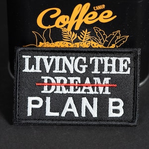 Könnte beinhalten: Ein rechteckiger, schwarz bestickter Aufnäher mit dem weißen Text LIVING THE, einer roten horizontalen Linie, die das Wort DREAM durchstreicht, und dem weißen Text PLAN B darunter. Der Aufnäher hat einen schwarzen Rand.