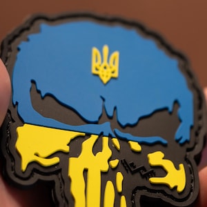 May include: A black patch featuring a skull design with the colors of the Ukrainian flag. The top half is blue, the bottom half is yellow, and a yellow trident is in the center.