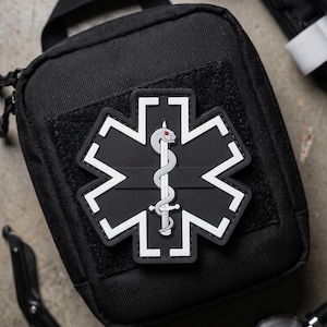 Pode incluir: Uma bolsa tática preta com um emblema da Estrela da Vida. O emblema é uma estrela preta de seis pontas com borda branca, centrada com uma espada prateada envolta por uma cobra cinza com um olho vermelho, fixada em um painel de velcro.