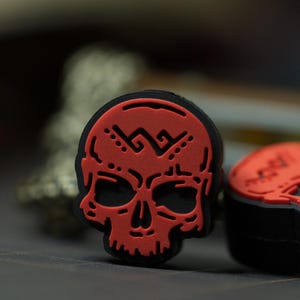 Puede incluir: Encanto de calavera roja con contorno negro y un dise&ntilde;o geom&eacute;trico en la frente. La calavera es un dise&ntilde;o en 3D y tiene una cara detallada.