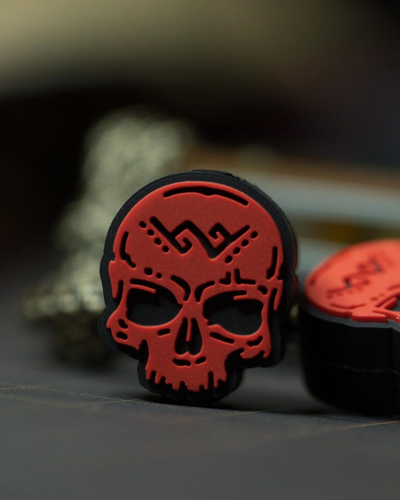 Puede incluir: Encanto de calavera roja con contorno negro y un dise&ntilde;o geom&eacute;trico en la frente. La calavera es un dise&ntilde;o en 3D y tiene una cara detallada.