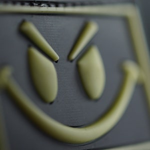 May include: A black patch with a yellow smiley face with a mischievous expression. The eyes are squinted and the mouth is curved into a smirk.