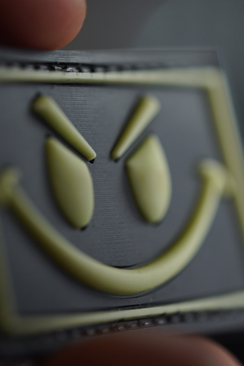May include: A black patch with a yellow smiley face with a mischievous expression. The eyes are squinted and the mouth is curved into a smirk.