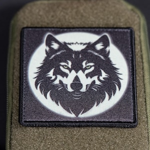 Puede incluir: Un parche cuadrado que presenta una ilustración gráfica en blanco y negro de una cabeza de lobo centrada sobre un fondo circular. El parche está sujeto a una superficie de tela verde oliva con un borde cosido visible alrededor del diseño.