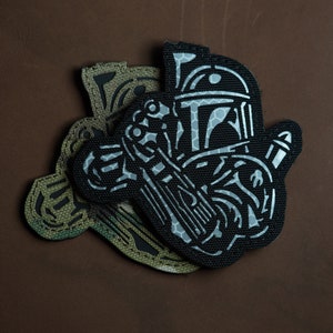May include: Two black and green embroidered patches, one featuring a stylized image of a Star Wars character, Boba Fett, with a rifle, and the other featuring a stylized image of a skull.