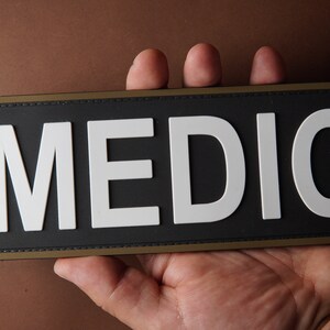 Pu&ograve; includere: Un cerotto nero con un bordo bianco e la parola "MEDIC" in lettere bianche.