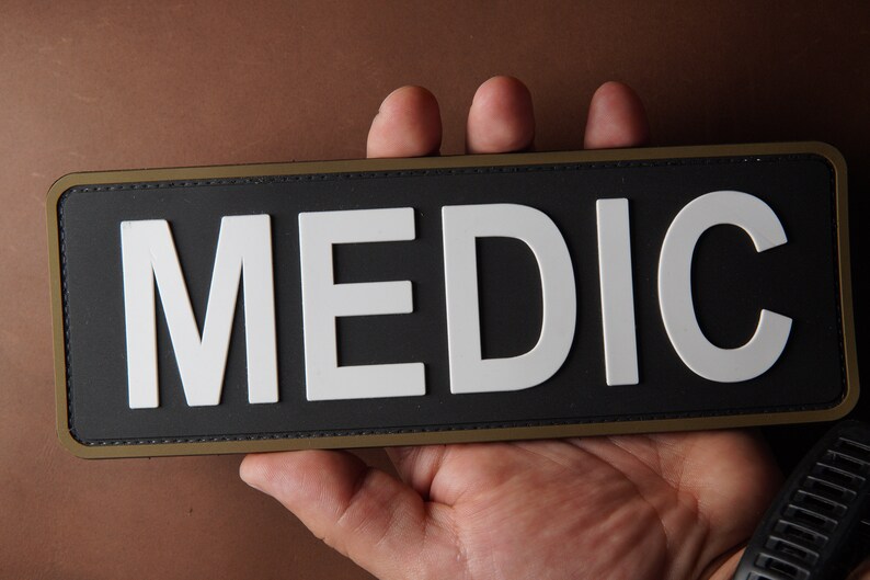Pu&ograve; includere: Un cerotto nero con un bordo bianco e la parola "MEDIC" in lettere bianche.
