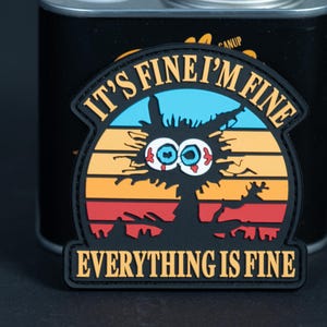 Könnte beinhalten: Ein schwarzer Gummi-Aufnäher mit einer Cartoon-Katze mit weit aufgerissenen, blutunterlaufenen Augen vor einem Sonnenuntergangshintergrund aus blauen, gelben, orangen und roten Streifen. Der Text IT'S FINE I'M FINE steht oben und unten.