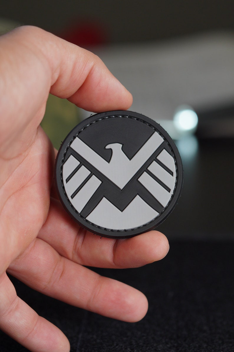 Puede incluir: Un parche circular negro y gris con un emblema de &aacute;guila estilizada. El dise&ntilde;o consiste en una silueta de &aacute;guila gris claro centrada sobre un fondo circular negro, con un borde cosido alrededor del contorno exterior del parche.