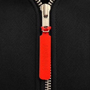 May include: A black ribbed fabric garment featuring a silver metal YKK zip closure. Attached to the zip pull is a long, rectangular, bright orange rubberised tag with the word JASMIN embossed vertically in capital letters.