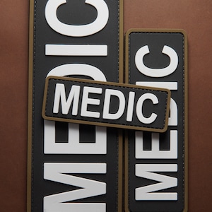 Könnte beinhalten: Zwei schwarze und weiße "MEDIC"-Patches mit olivgrünen Nähten. Die Patches sind rechteckig und haben ein erhabenes, strukturiertes Design.