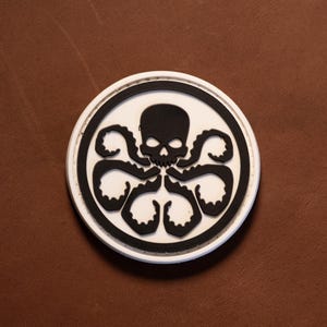 May include: A circular patch featuring a black skull and octopus tentacles centered on a white background, enclosed by a thick black border and a thin white outer rim. The design is flat and graphic, displaying a stylized cephalopod creature.