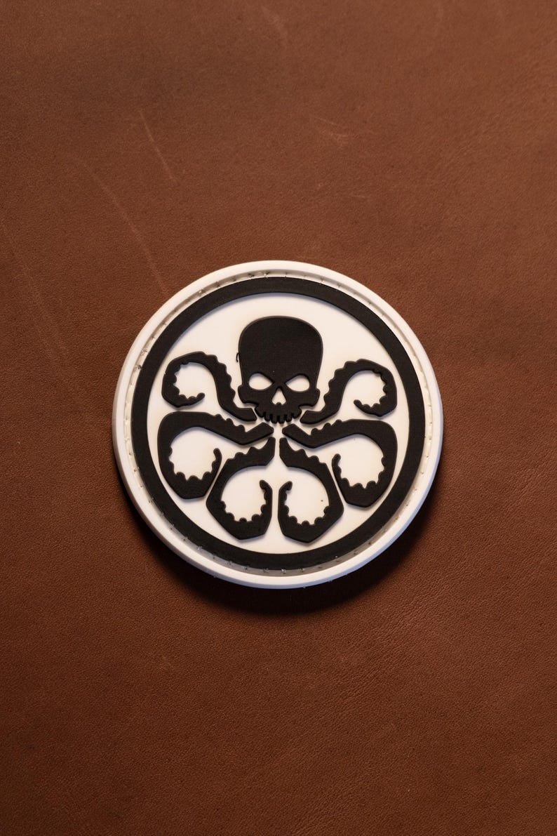 May include: A circular patch featuring a black skull and octopus tentacles centered on a white background, enclosed by a thick black border and a thin white outer rim. The design is flat and graphic, displaying a stylized cephalopod creature.