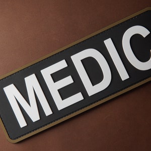 Pu&ograve; includere: Un cerotto nero con un bordo bianco e la parola "MEDIC" in lettere bianche.