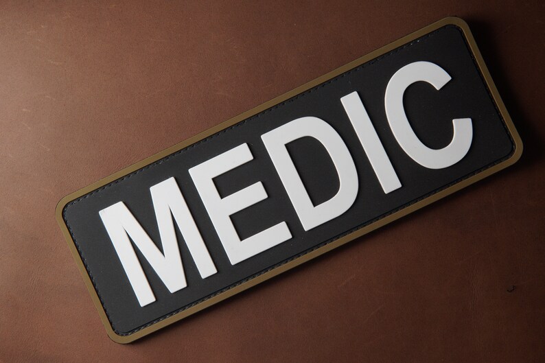 Pu&ograve; includere: Un cerotto nero con un bordo bianco e la parola "MEDIC" in lettere bianche.