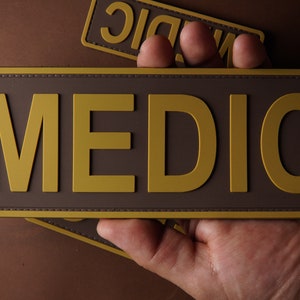Pu&ograve; includere: Una toppa marrone con un'etichetta dorata "MEDIC". La toppa ha un bordo cucito.