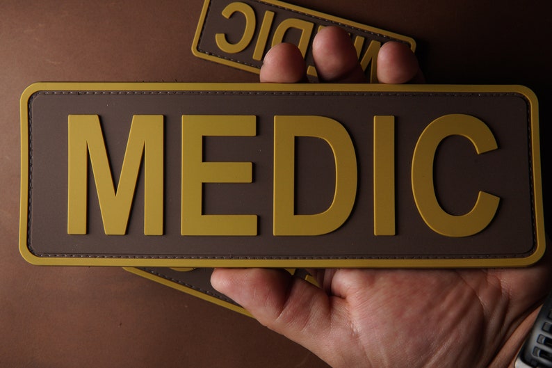 Pu&ograve; includere: Una toppa marrone con un'etichetta dorata "MEDIC". La toppa ha un bordo cucito.