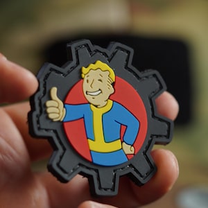 May include: A black and red patch with a cartoon character wearing a blue jumpsuit and a yellow belt. The character is giving a thumbs up. The patch is shaped like a gear.