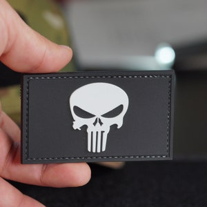 Pode incluir: Um remendo preto com um design de caveira e ossos cruzados brancos. A caveira tem uma expressão ameaçadora com a testa franzida e a testa enrugada. O remendo é feito de um material macio e flexível e tem uma borda costurada.